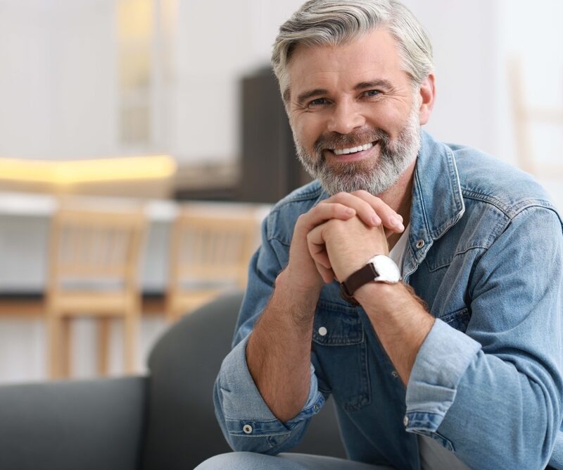 Smiling man with a gray beard in a denim shirt on a sofa, reflecting the confidence of tooth-colored fillings in La Mesa, CA