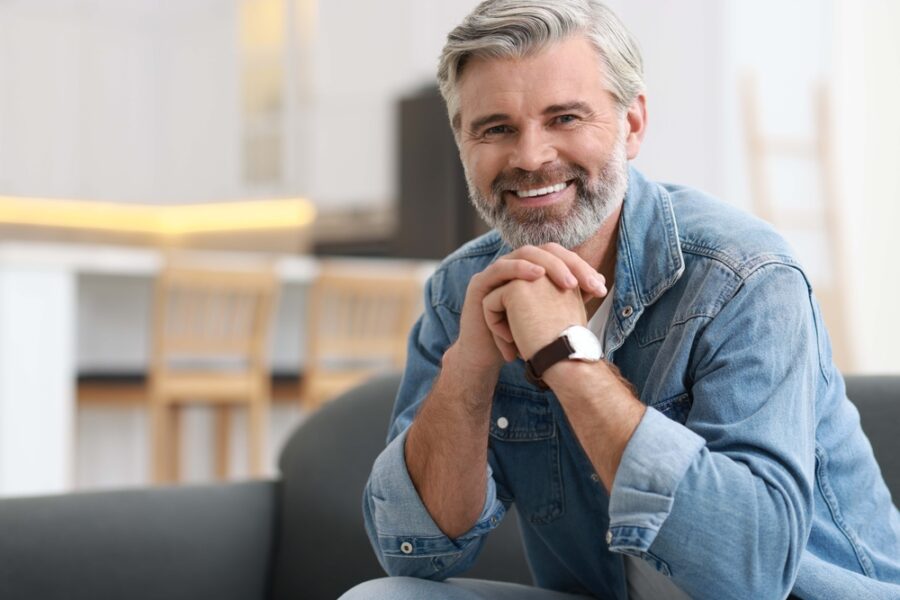 Smiling man with a gray beard in a denim shirt on a sofa, reflecting the confidence of tooth-colored fillings in La Mesa, CA