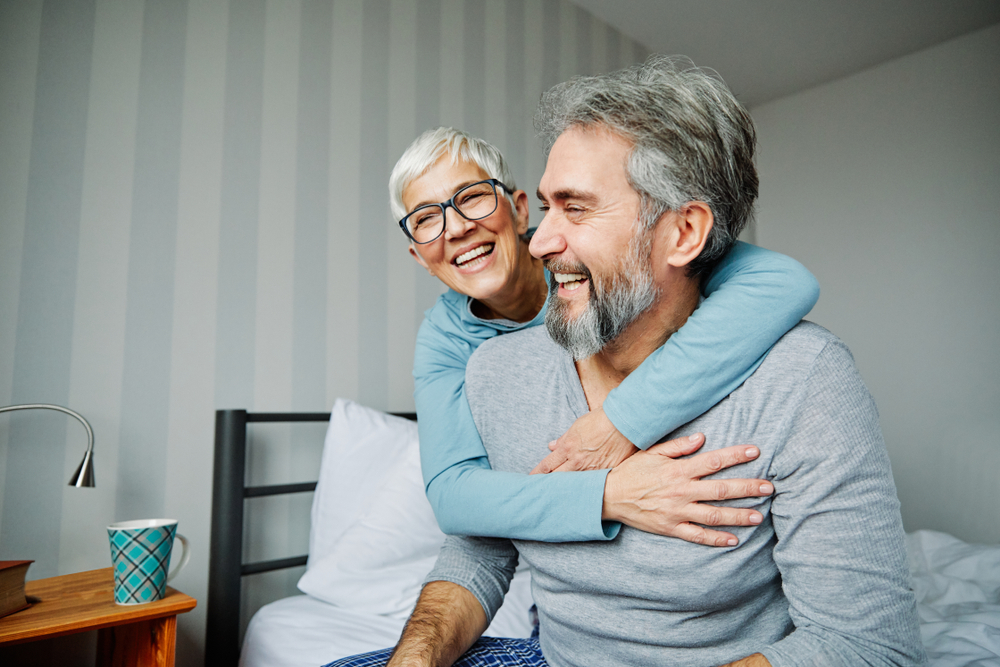 Laughing senior couple hugging on a bed, reflecting the relief of prompt emergency dental care in La Mesa, CA