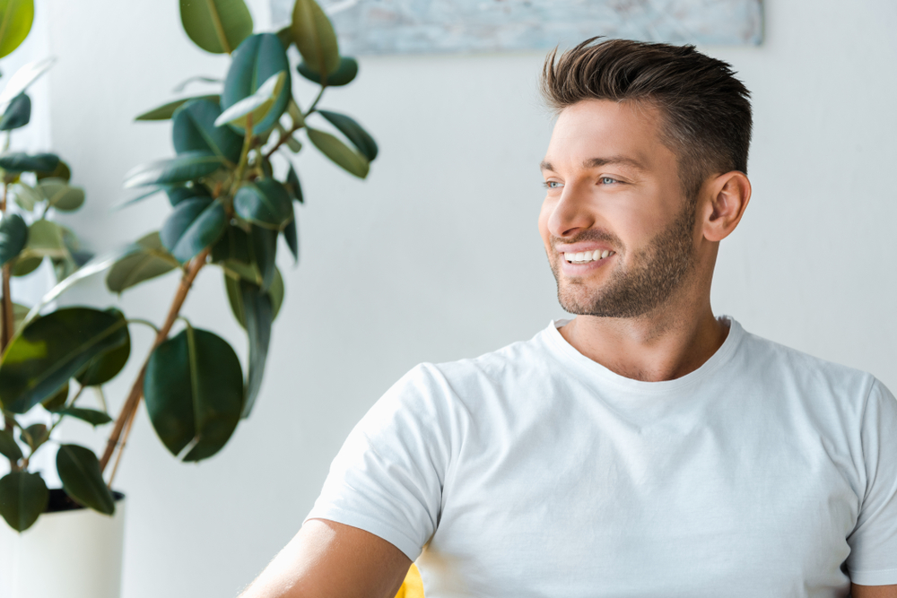 Confident Smile at Home, Dental Veneers in La Mesa, CA Smiling man with a trimmed beard in a white T-shirt beside a houseplant, reflecting the confidence of veneers in La Mesa, CA