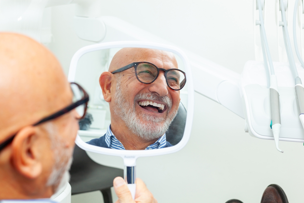 Implant Dentist in La Mesa, CA: Smile Check in the Mirror Bearded senior man in glasses laughing at his smile in a handheld mirror at an implant dentistry office in La Mesa, CA
