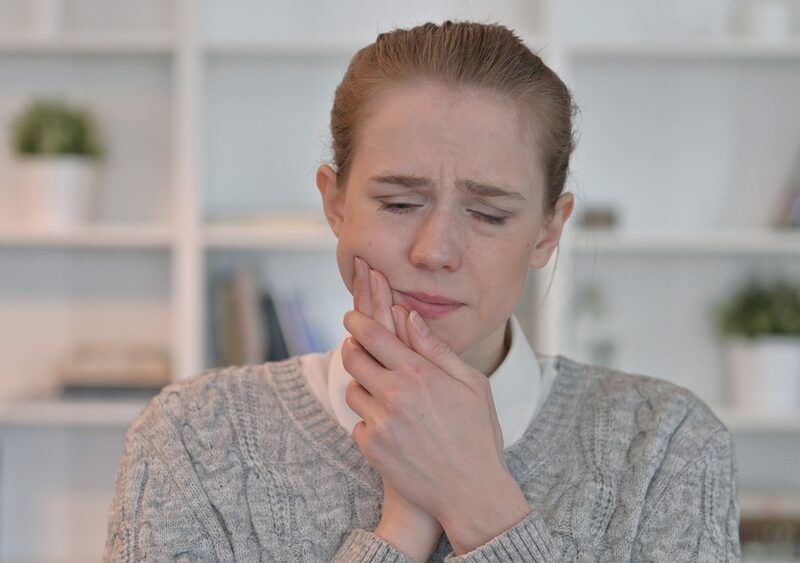 Woman in a gray sweater wincing and holding her jaw from a toothache, a sign to see an emergency dentist in La Mesa, CA