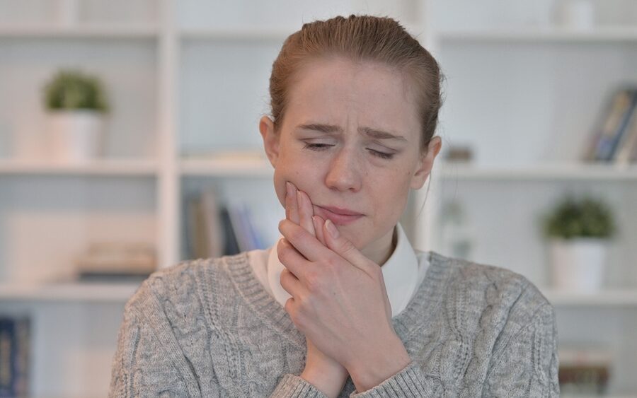 Woman in a gray sweater wincing and holding her jaw from a toothache, a sign to see an emergency dentist in La Mesa, CA