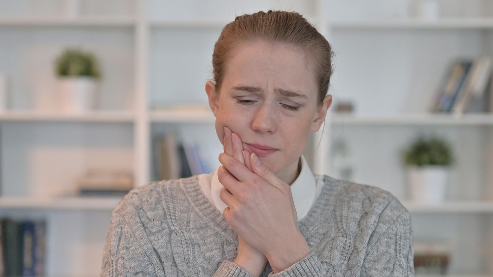 Woman in a gray sweater wincing and holding her jaw from a toothache, a sign to see an emergency dentist in La Mesa, CA