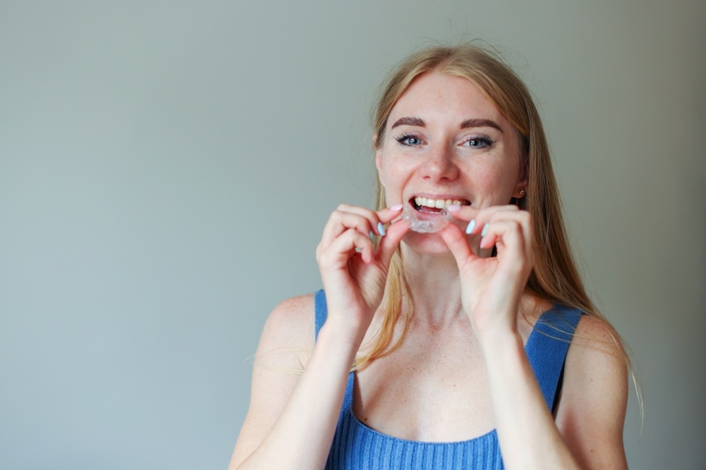 Invisalign in La Mesa, CA: Wearing Clear Aligners Smiling woman in a blue tank top placing a clear aligner over her teeth, illustrating Invisalign treatment in La Mesa, CA