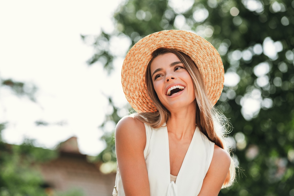 Laughing woman in a straw hat and white top outdoors, reflecting the comfort of timely urgent dental care in La Mesa, CA